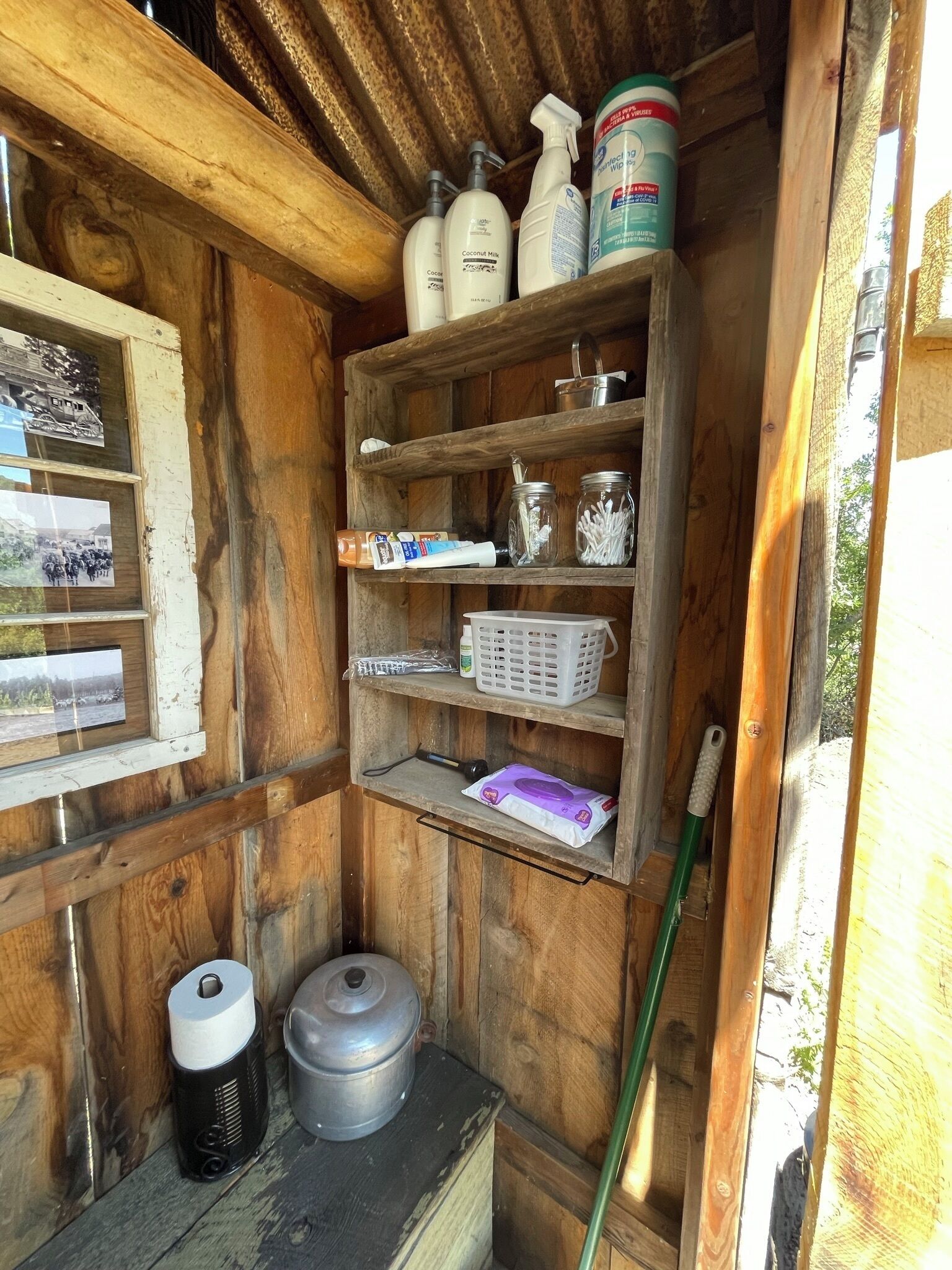 A rustic wooden cabin interior with shelves holding cleaning supplies, a window with photos, and a small storage area with a metal container and a broom.