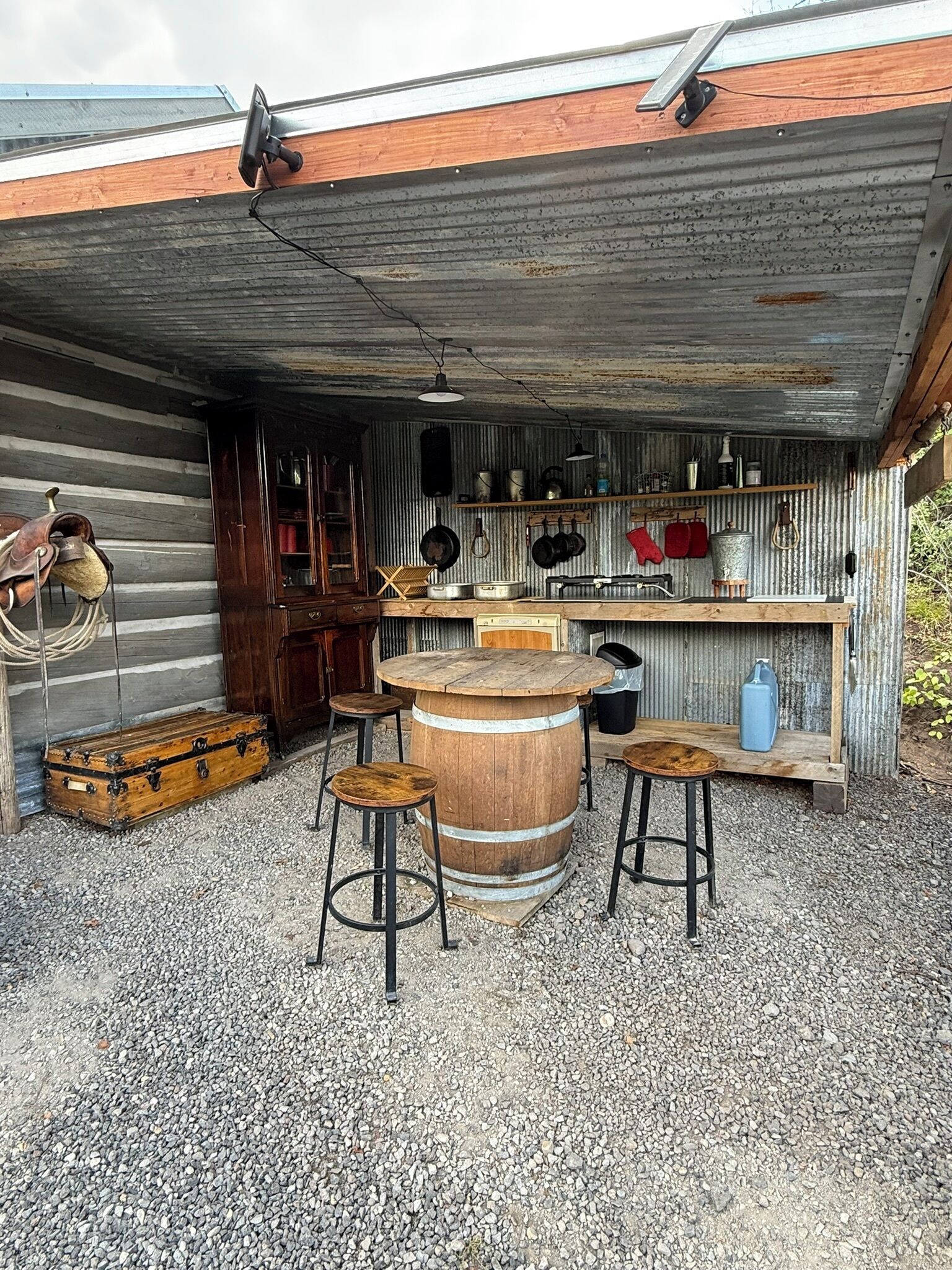 A rustic outdoor kitchen with a wooden barrel table, stools, and a metal sink.