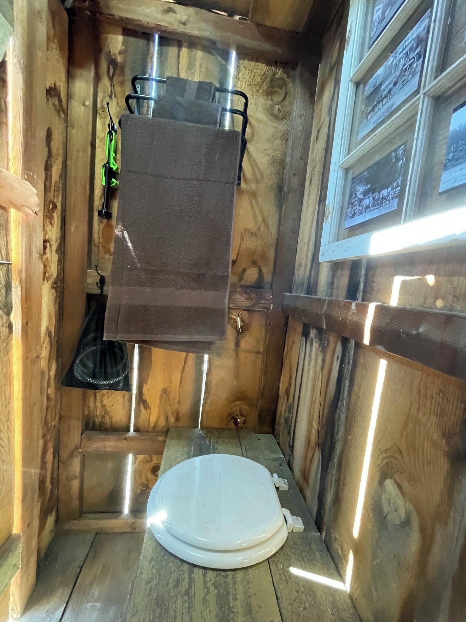 A compact wooden bathroom with a white toilet, a hanging towel rack, and a window with a view.