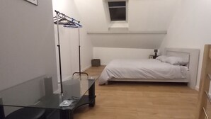 Un dormitorio pequeño y bien iluminado con una cama individual, piso de madera, un escritorio de vidrio y una ventana sobre la cama.