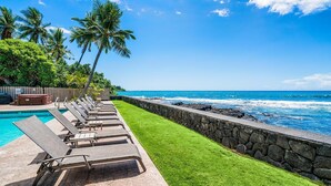 A poolside area with lounge chairs, a stone wall, and a view of the ocean. - Banyan Tree 203 - Peaceful oceanfront Kona condo (Kailua Kona)
