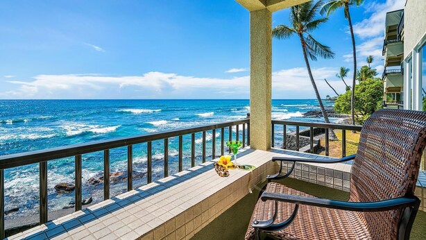 A balcony with a view of the ocean, a chair, a table with a drink, and a tropical plant. - Banyan Tree 203 - Peaceful oceanfront Kona condo (Kailua Kona)