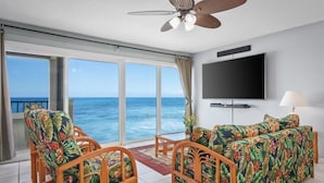 A room with a large window overlooking the ocean, a ceiling fan, a flat-screen TV, and a sofa with floral upholstery. - Banyan Tree 203 - Peaceful oceanfront Kona condo (Kailua Kona)