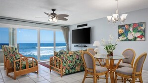A room with a large window overlooking the ocean, a ceiling fan, a flat-screen TV, a dining table with chairs, and a floral sofa. - Banyan Tree 203 - Peaceful oceanfront Kona condo (Kailua Kona)