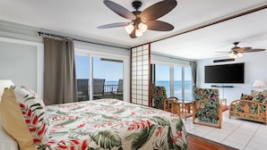 A bedroom with a bed, a TV, a ceiling fan, a view of the ocean, and a balcony. - Banyan Tree 203 - Peaceful oceanfront Kona condo (Kailua Kona)