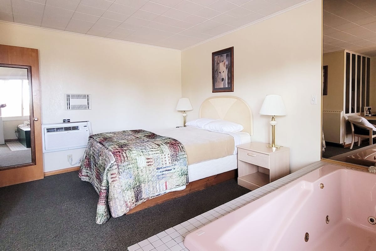 A hotel room with a bed, two bedside tables with lamps, a large window, a wall-mounted air conditioning unit, a bathroom with a bathtub, and a door leading to another room.