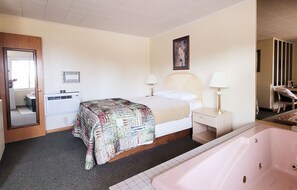 Habitación de hotel con cama, dos mesitas de noche con lámparas, un ventanal grande, unidad de aire acondicionado instalada en la pared, baño con bañera y una puerta que da a otra habitación.