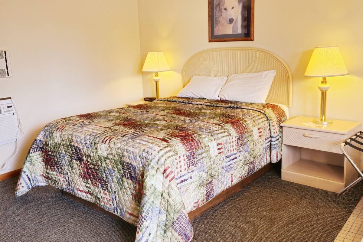 A neatly made bed with a colorful quilt, two bedside lamps, a framed picture on the wall, and a small bedside table with a lamp.