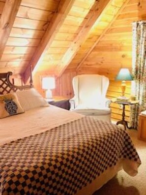A cozy attic bedroom with a wooden ceiling, a bed with a patterned bedspread, a chair, and a small table with a lamp.