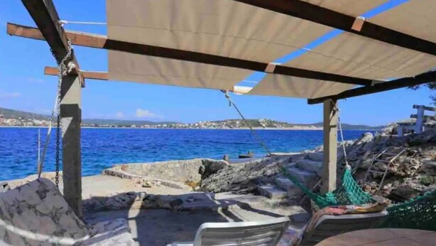 En terrasse ved havet med et bord, stole og en hængekøje, samt havudsigt og bygninger i det fjerne.