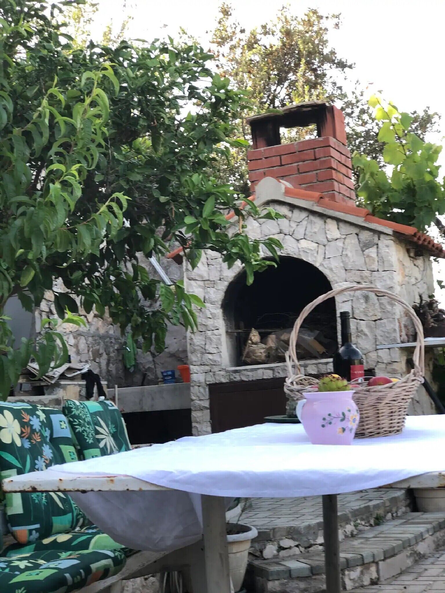 Un horno de piedra al aire libre con chimenea de ladrillo, rodeado de follaje y una mesa con una cesta de frutas y una botella de vino.