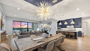 A spacious living area with a large wooden dining table, wicker chairs, a modern chandelier, and a view of the outdoors through large windows.