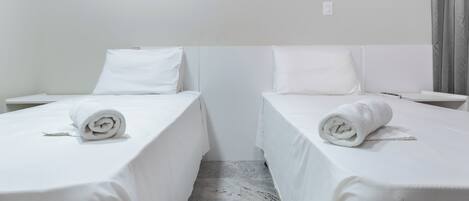A hotel room with two single beds, each with a rolled white towel. The room has white bedding and a marble floor.