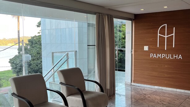 En modern hotellobby med två beige fåtöljer, en väggpanel i trä med hotellets namn och utsikt mot utomhusmiljön.