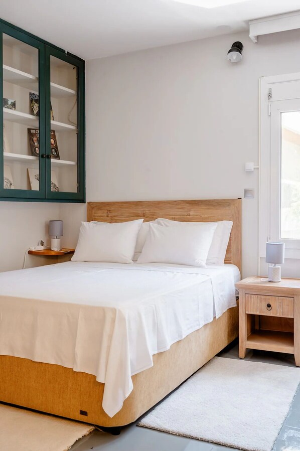 Une chambre à coucher avec une tête de lit en bois, du linge de lit blanc, une table de chevet avec une lampe et une armoire à cadres verts contenant des livres.