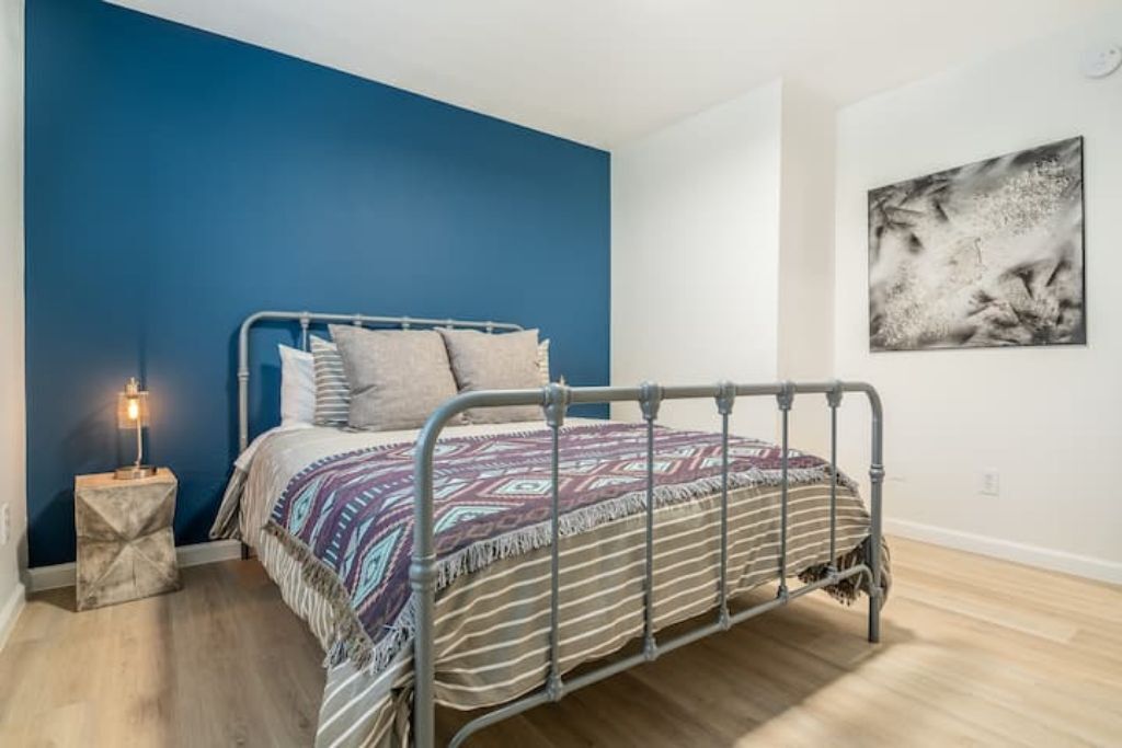 A bedroom with a metal bed frame, a patterned bedspread, a bedside lamp, and a wall-mounted abstract painting.