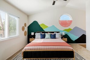 A bedroom with a bed, a window with blinds, a wall mural, and a round mirror.