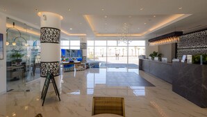 A modern hotel lobby with a reception desk, a large chandelier, and a glass wall with a view of palm trees. - Encantame Towers (Puerto Peñasco)