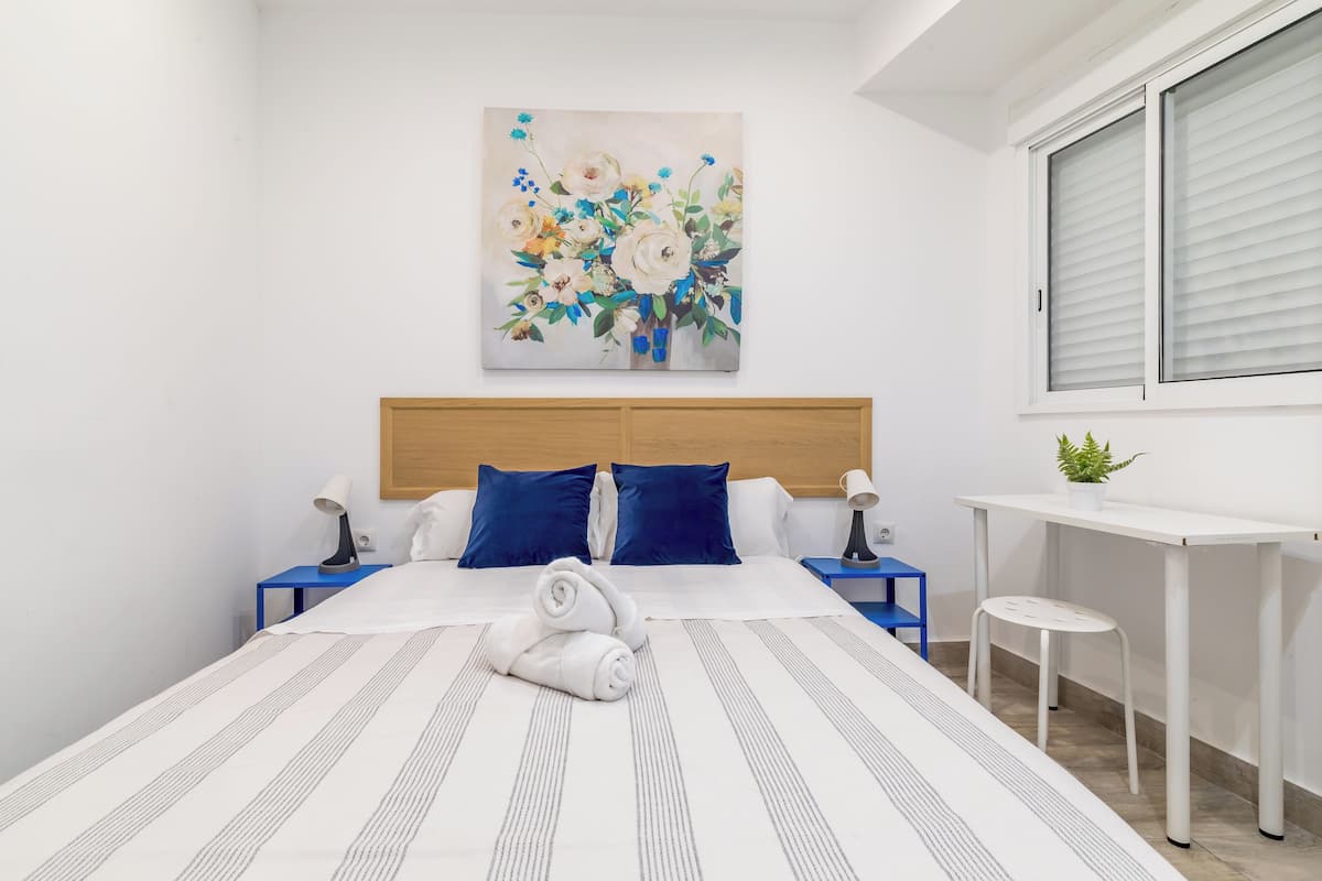 A neatly made bed with a striped bedspread, a painting of flowers on the wall, a desk with a chair, and a window with blinds.