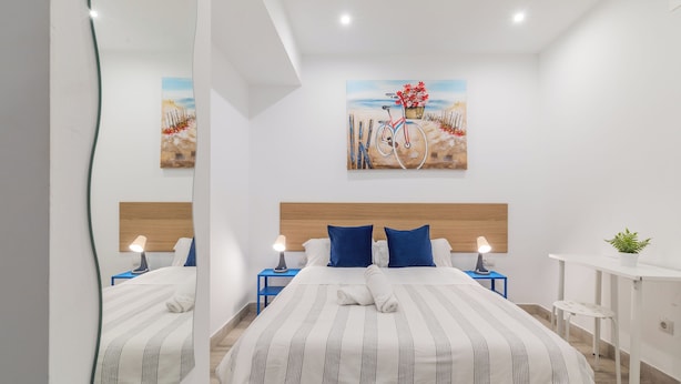 A modern hotel room with a large bed, a nightstand with a lamp, a small desk with a plant, and a painting on the wall. - LooP Las Ventas - Adults Only (Madrid)