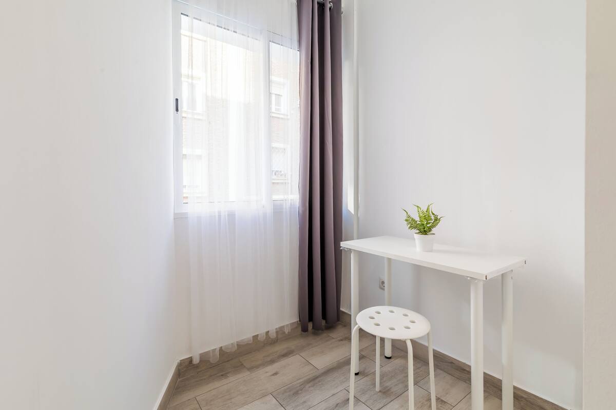 A small, bright room with a white table and a single chair, a potted plant on the table, and a window with sheer curtains.