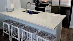 A modern kitchen with a large island, white bar stools, stainless steel appliances, and decorative wall art. - Hightide Hideaway Beach Getaway. (Corpus Christi)