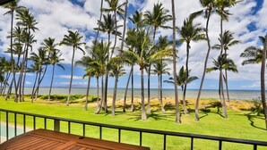 A balcony with a wooden table and lounge chairs overlooking a beach with palm trees and the ocean. - Oceanfront, Hale Kai O Kihei, incredible sunsets, King bed, AC, remodeled 2023 (Kihei)