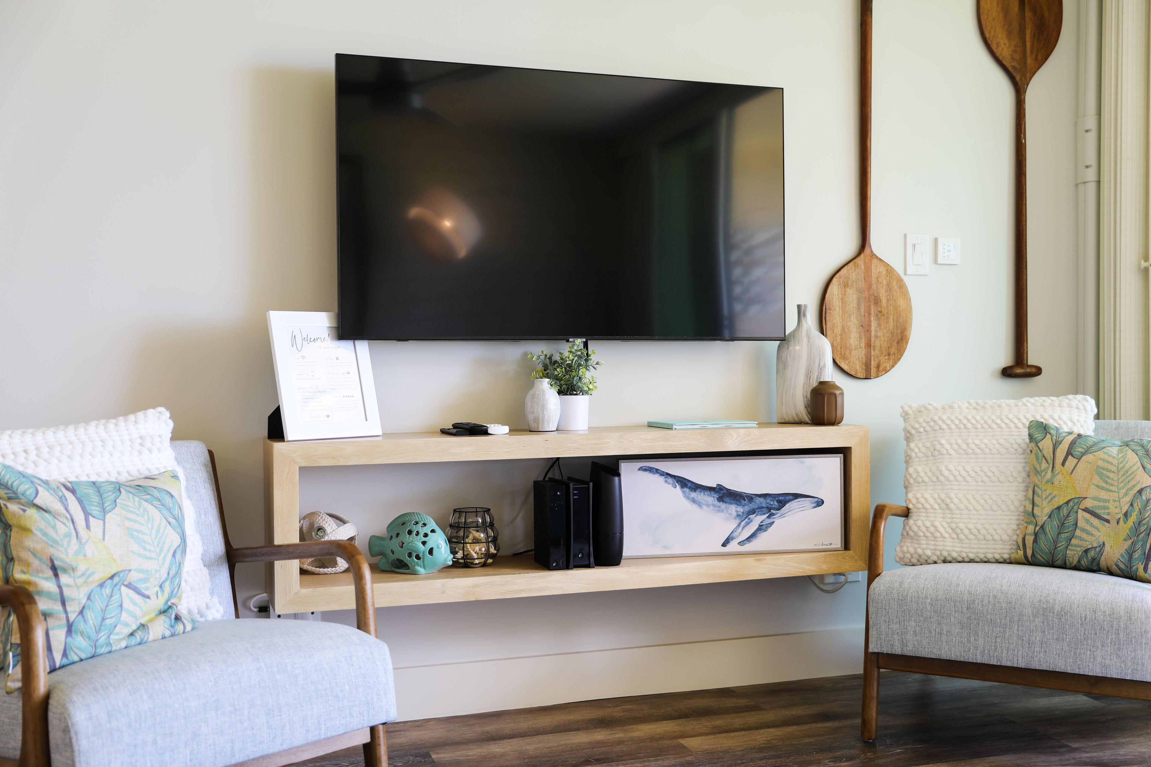 A living room with a flat-screen TV mounted on the wall, wooden furniture, a shelf with decorative items, and a framed picture of a whale.