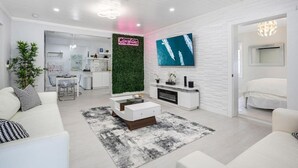 A modern living room with a white sofa, a coffee table, and a TV mounted on a white brick wall. There is a kitchen visible in the background.