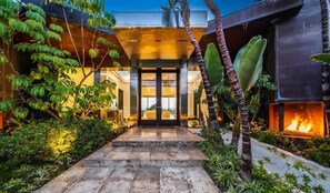 Une maison moderne avec un chemin de pierre menant à une porte en verre, entourée d’une végétation luxuriante et agrémentée d’un foyer.