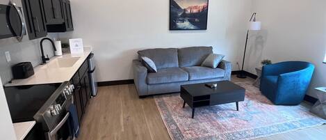 A modern kitchen with dark cabinets, a built-in oven, and a sink. A gray sofa and a black coffee table are in the living area, with a colorful rug on the floor.