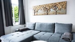 A modern living room with a grey sectional sofa, a white coffee table, and a painting of trees on the wall. - Wunderschöne Ferienwohnung Inmitten der Altstadt (Schwerin)