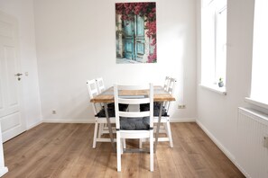A dining area with a wooden table and white chairs, a painting of doors above, and a window with a plant. - Wunderschöne Ferienwohnung Inmitten der Altstadt (Schwerin)