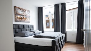 A modern bedroom with a large bed, a headboard, a window with curtains, and a wall-mounted artwork. - Wunderschöne Ferienwohnung Inmitten der Altstadt (Schwerin)