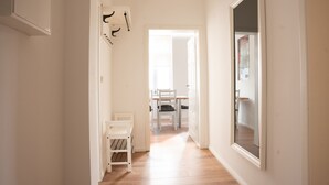 A hallway with a white door, a small white table, and a chair. There is a coat rack with several coats hanging on it. The floor is wooden, and the walls are white. - Wunderschöne Ferienwohnung Inmitten der Altstadt (Schwerin)