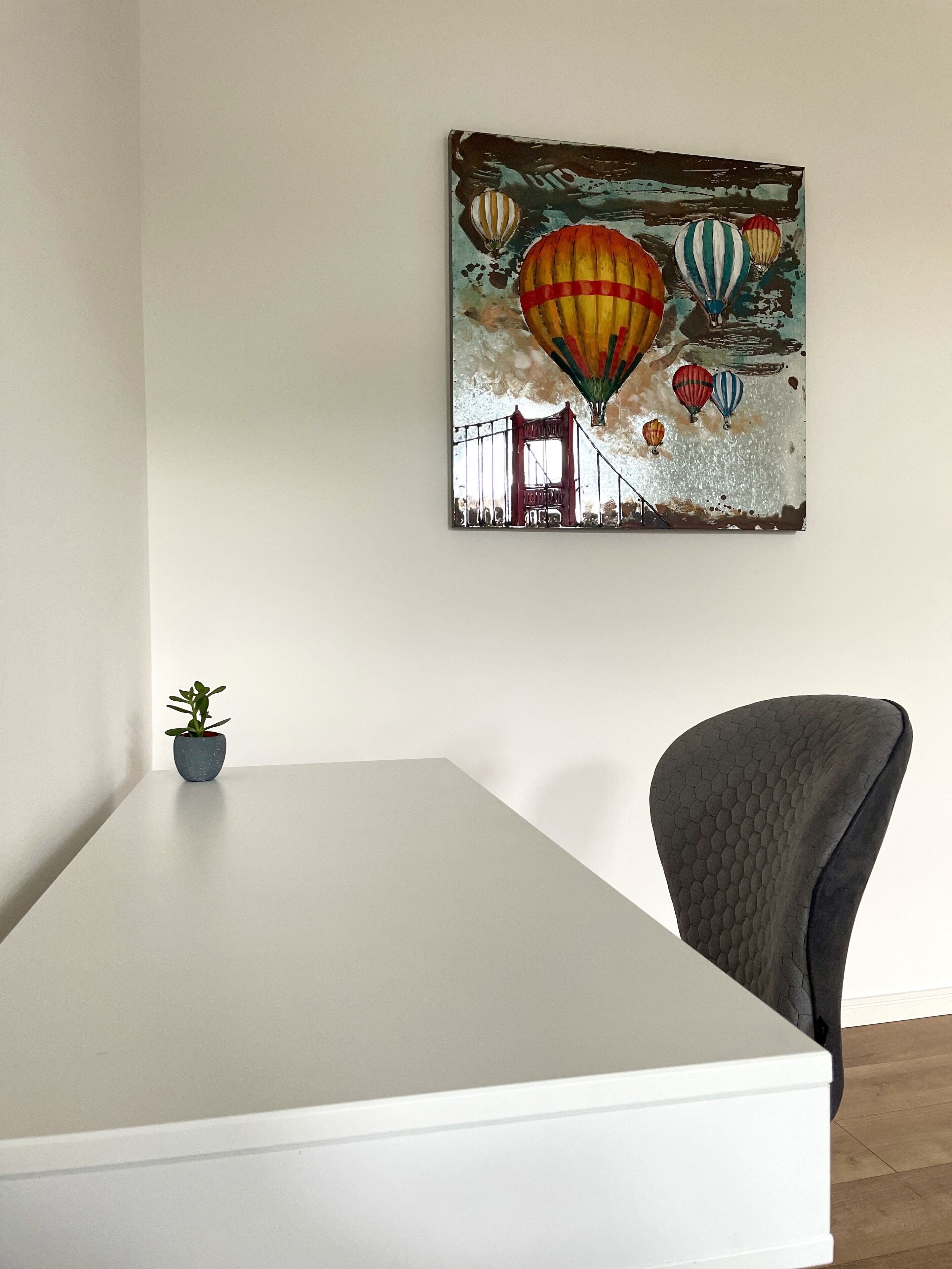 Une chambre moderne avec un bureau blanc, une chaise grise et un tableau représentant des montgolfières, accroché au mur.
