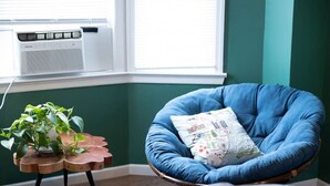A room with a blue chair, a small wooden table with a plant, and a window with an air conditioner.