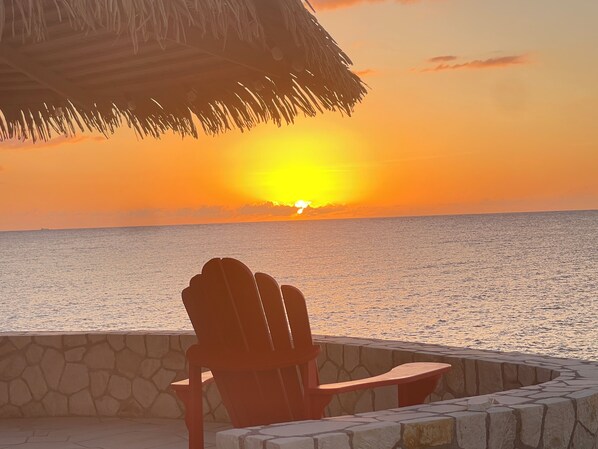 A sunset view from a stone-walled patio with a thatched roof, overlooking the ocean. - Oceanfront, breathtaking views of turquoise Caribbean Sea, gated w/private beach (Negril)