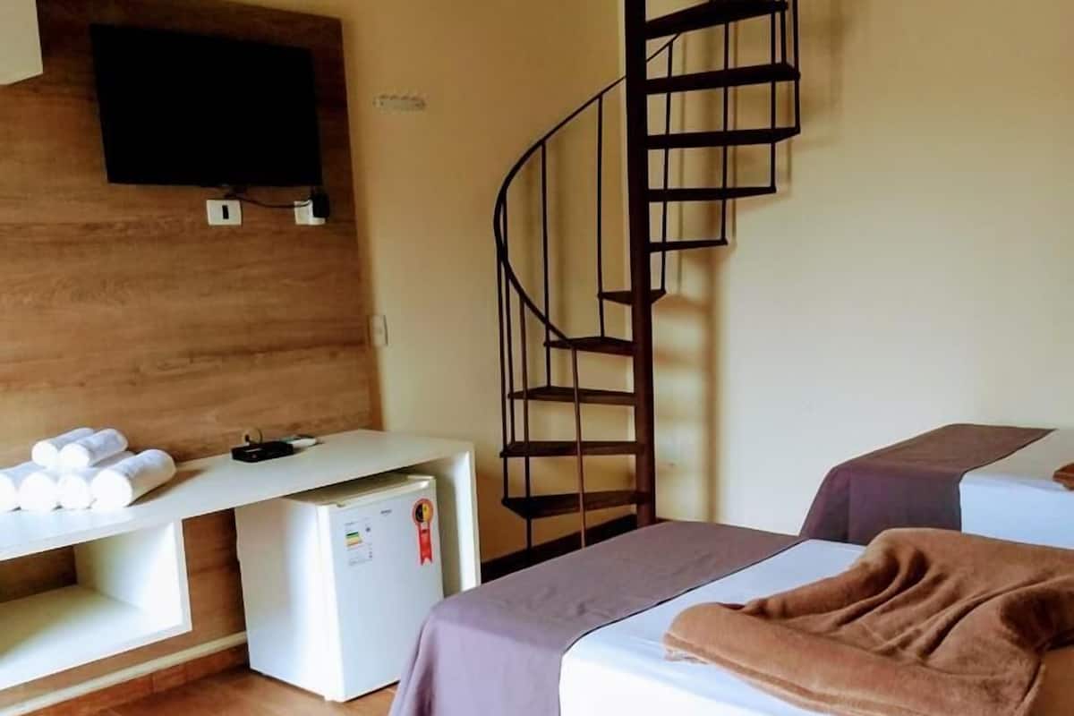 A bedroom with two beds, a small desk, a refrigerator, a TV mounted on the wall, and a spiral staircase.