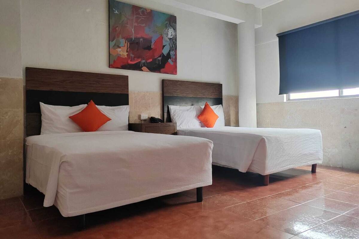 A hotel room with two beds, each with white bedding and orange pillows, a nightstand with a phone, and a large abstract painting on the wall.