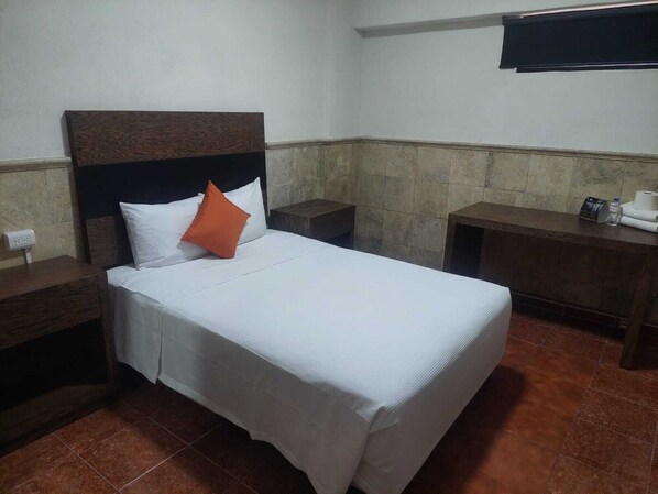 Una habitación de hotel con una cama, una mesita de noche, un cabecero de madera, un televisor montado en la pared y un piso de baldosas.