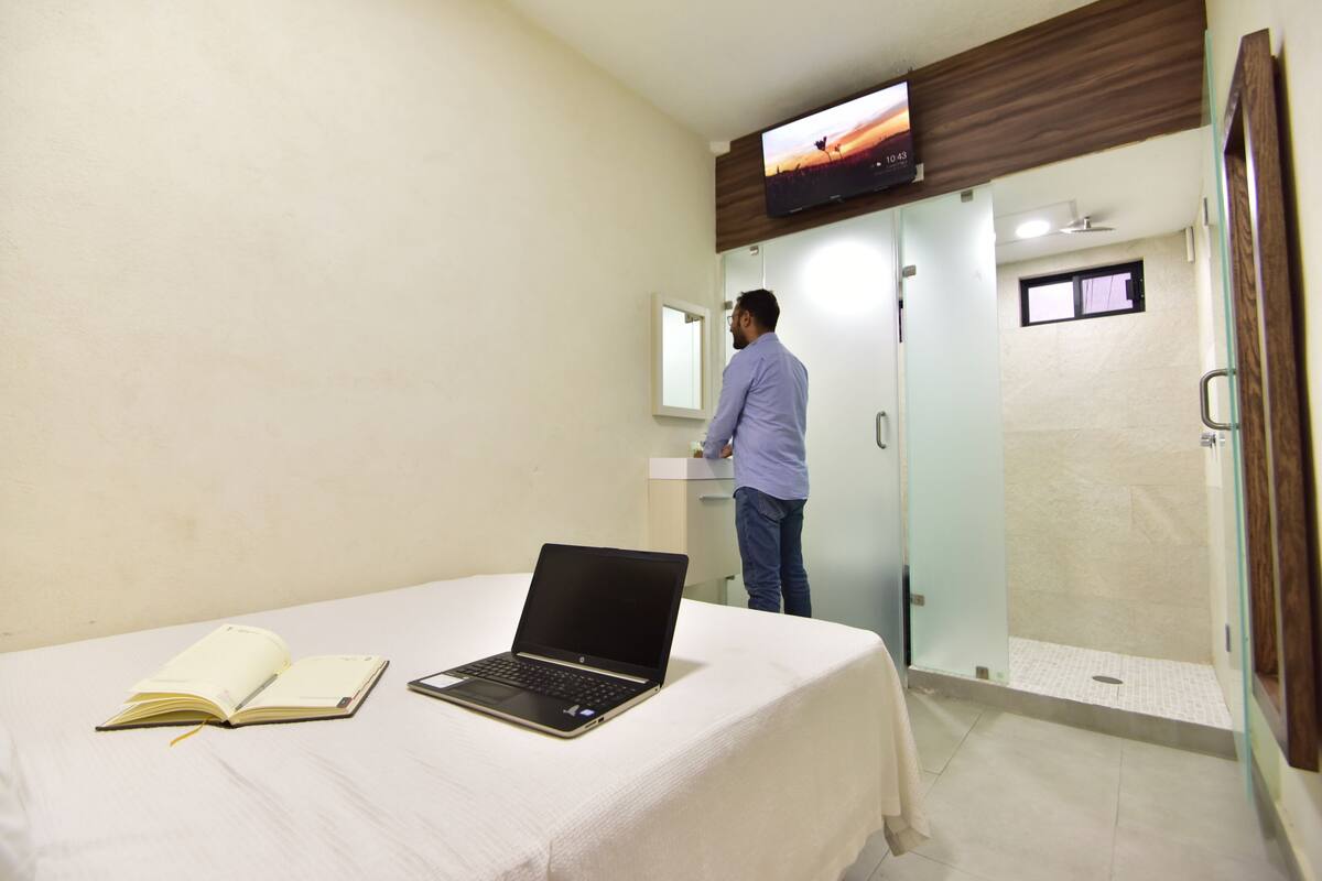 A hotel room with a bed, a laptop, an open book, a TV mounted on the wall, and a shower area.