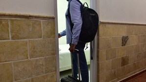 Una persona con maleta y mochila parada en un pasillo con piso de baldosas y una puerta que da a una habitación.