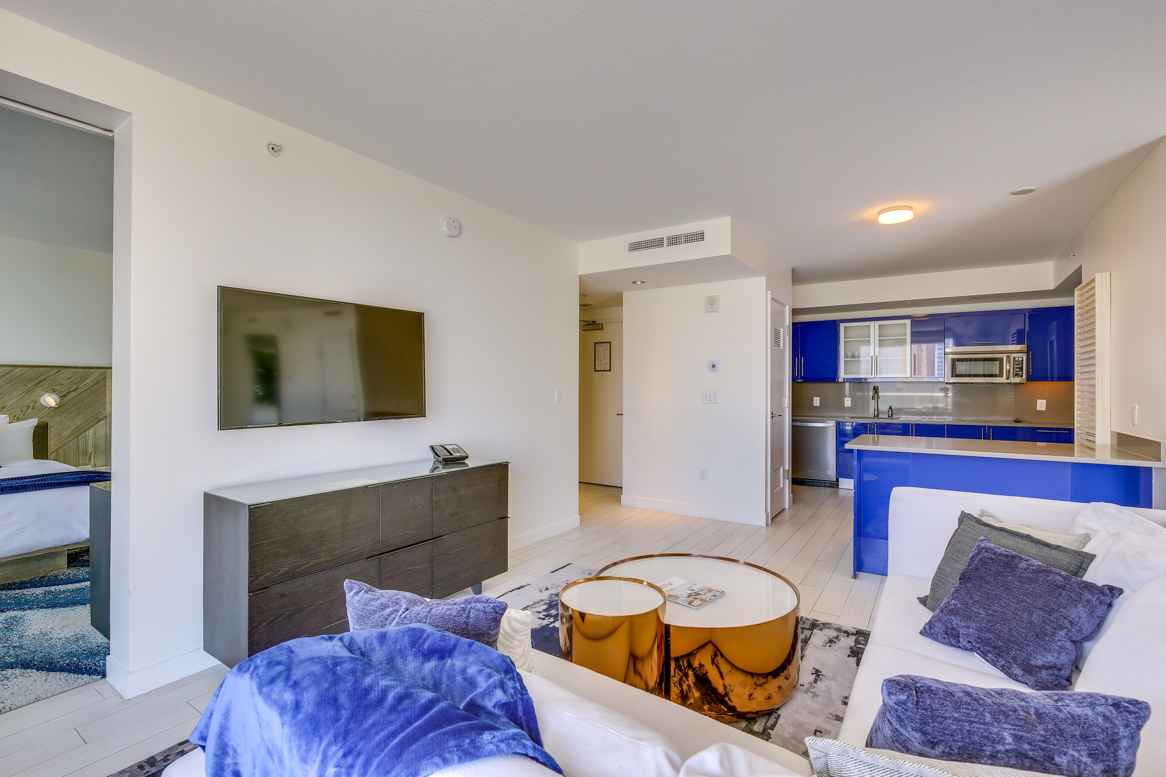 A modern hotel room with a sofa, a coffee table, a TV mounted on the wall, and a kitchenette in the background.
