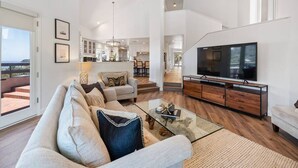 A spacious living room with a large sofa, a coffee table, and a television. There is a kitchen in the background with white cabinets and a dining area. - Mansion in La Jolla Heights-Pool, Spa, OceanViews (San Diego)