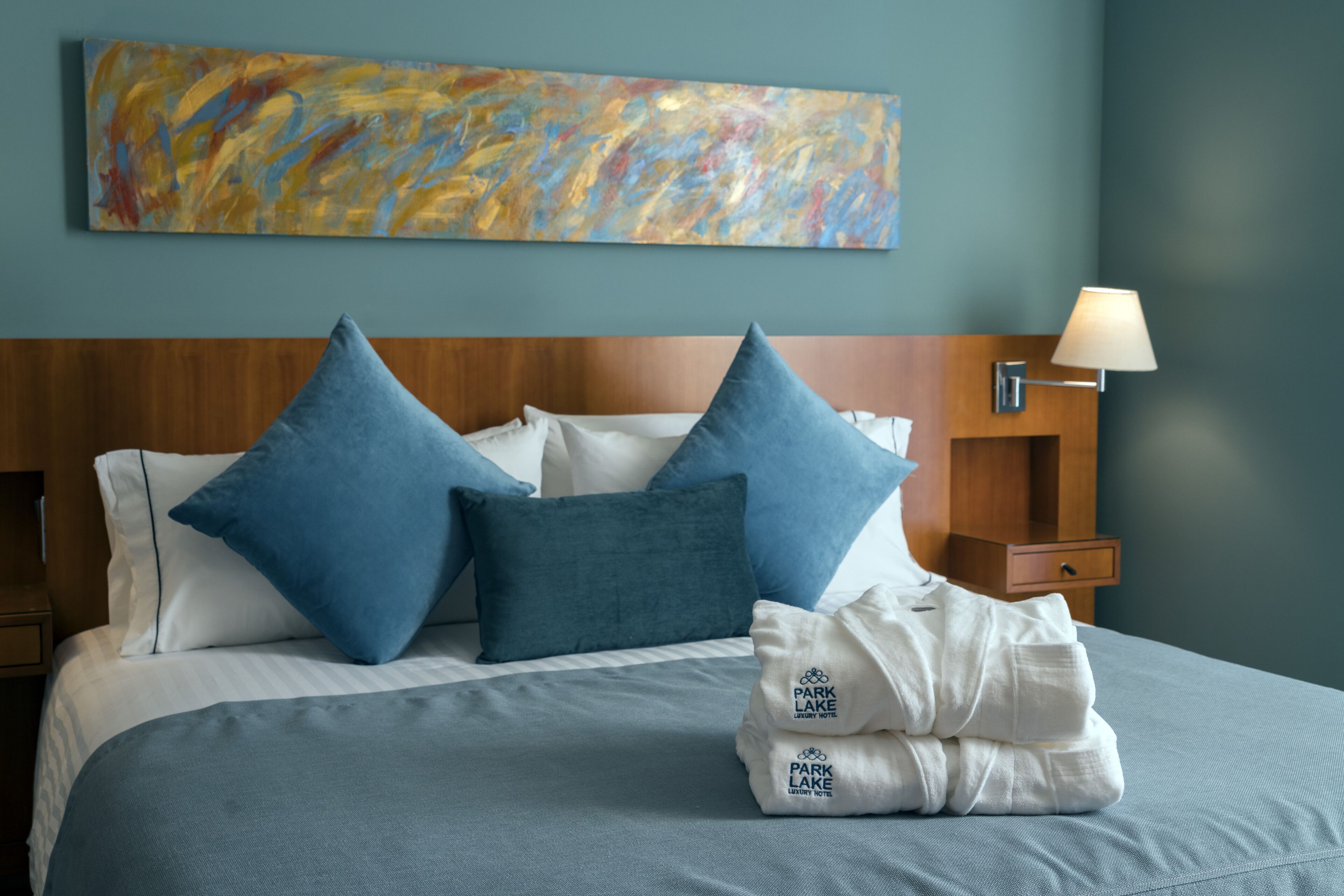 A neatly made bed with blue and white pillows, a blue blanket, and a white towel with "Park Lake Luxury Hotel" branding.