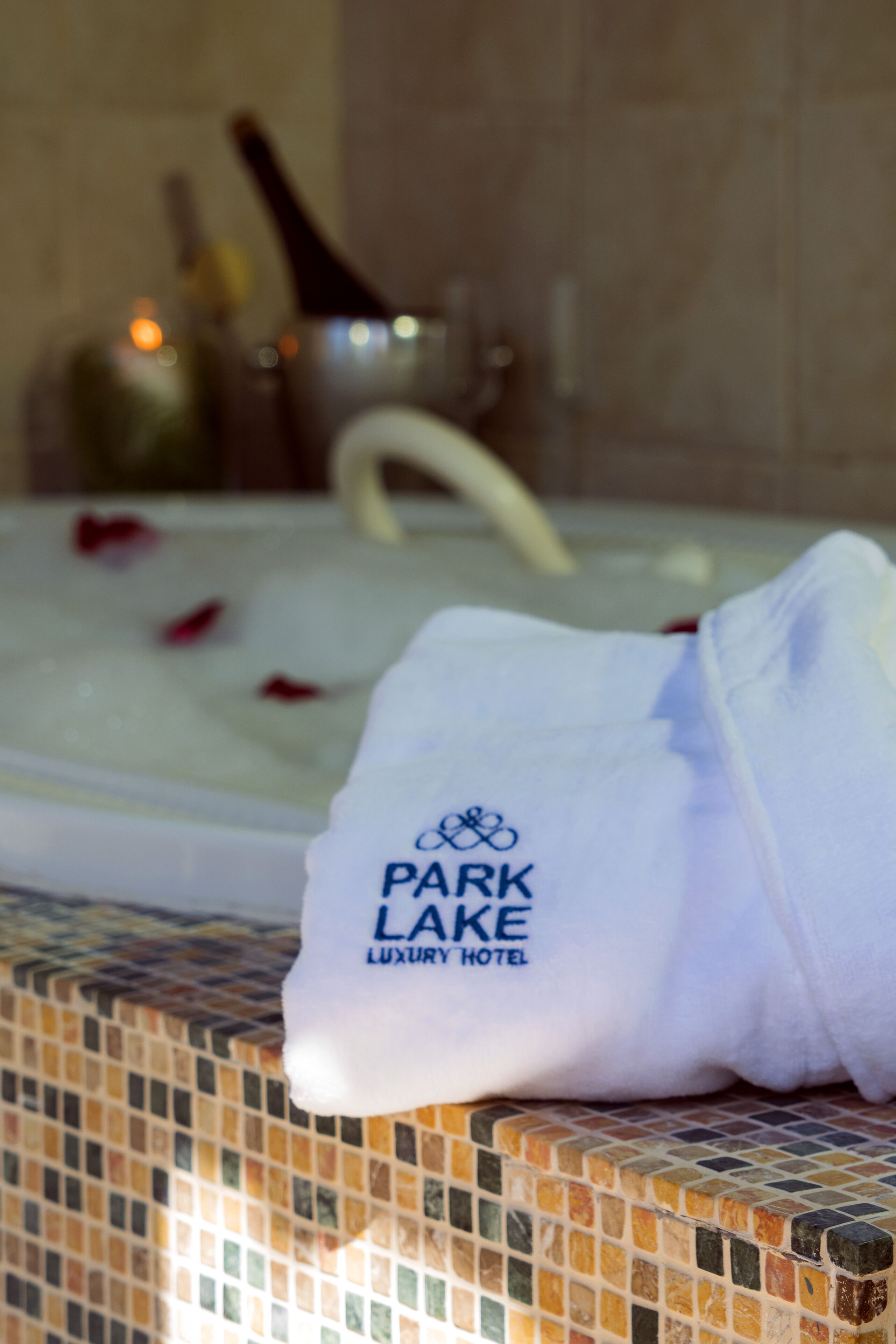 A white towel with "Park Lake Luxury Hotel" logo on a tiled surface near a bathtub with rose petals.