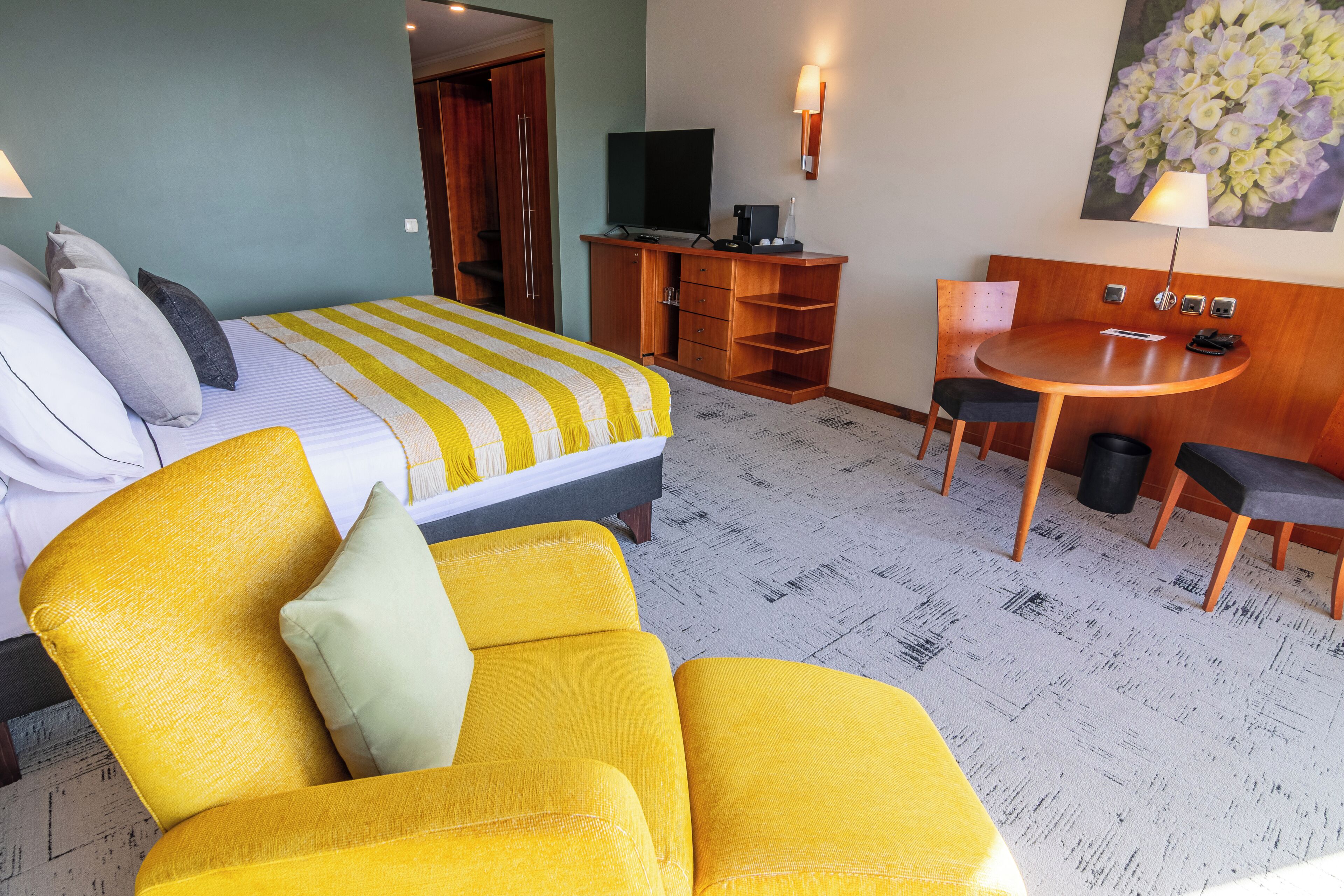 Ein Hotelzimmer mit einem Bett, einem gelben Sessel, einem kleinen Tisch mit Stühlen, einem Schreibtisch mit Fernseher und einem Bild an der Wand.