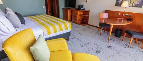A hotel room with a bed, a yellow armchair, a small table with chairs, a desk with a TV, and a painting on the wall.