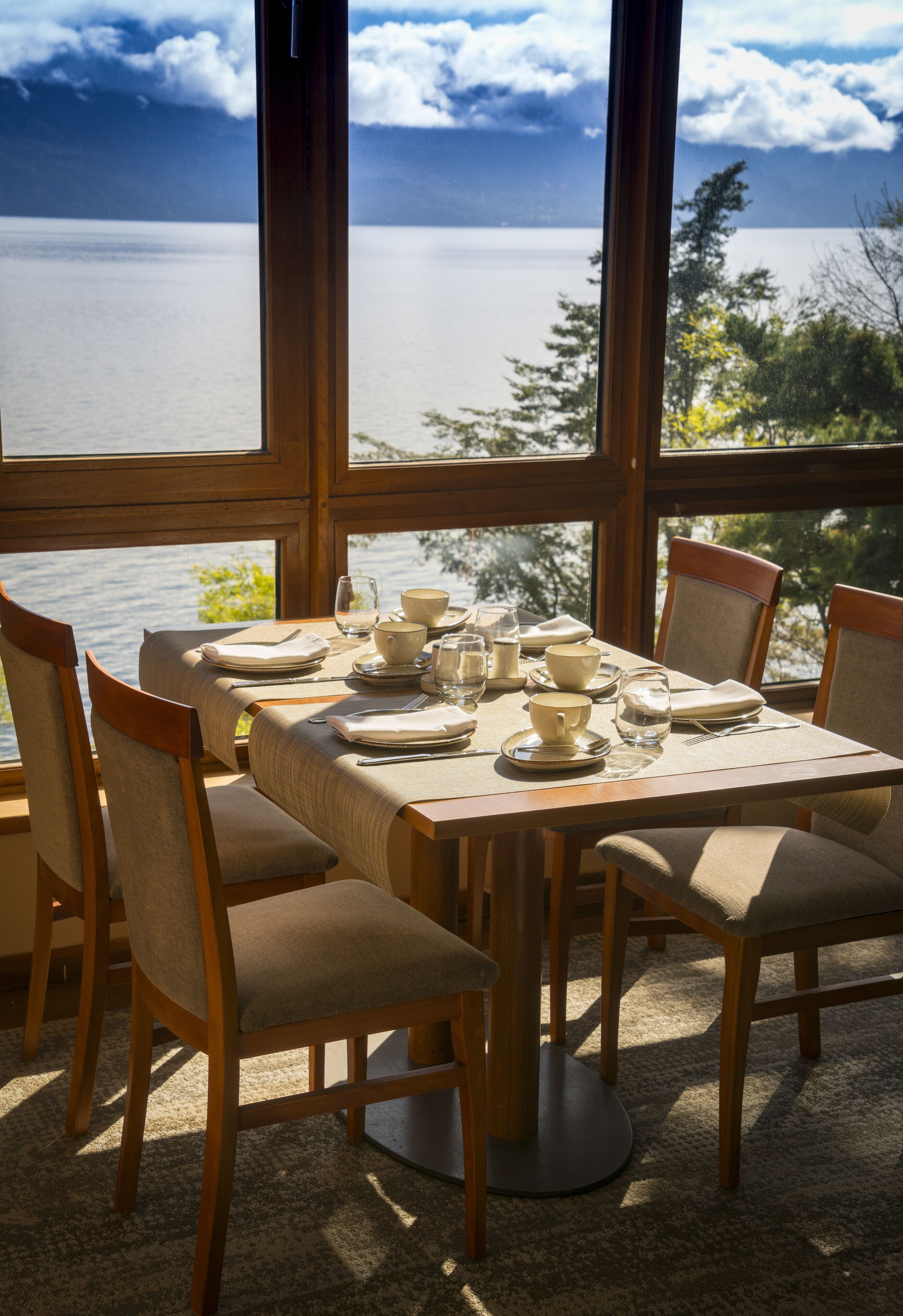A dining table set for four with chairs, positioned in a room with large windows overlooking a body of water.